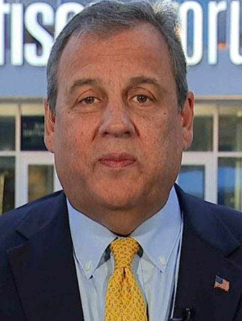 Man in a dark suit and yellow tie standing in front of a building.