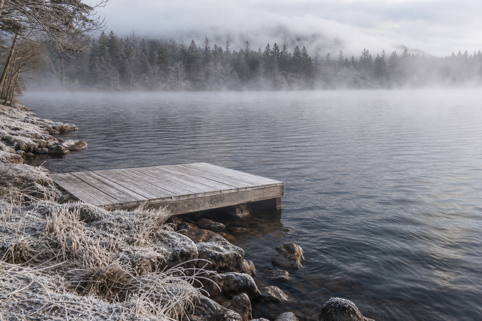Wooden dock beside a foggy lake with misty mountains and frosty shoreline