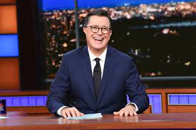 Smiling late-night host seated at a desk in a studio with city lights behind him.