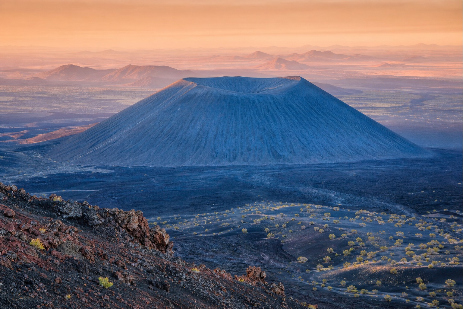 A blue cinder cone rising above a vast desert landscape at sunset.