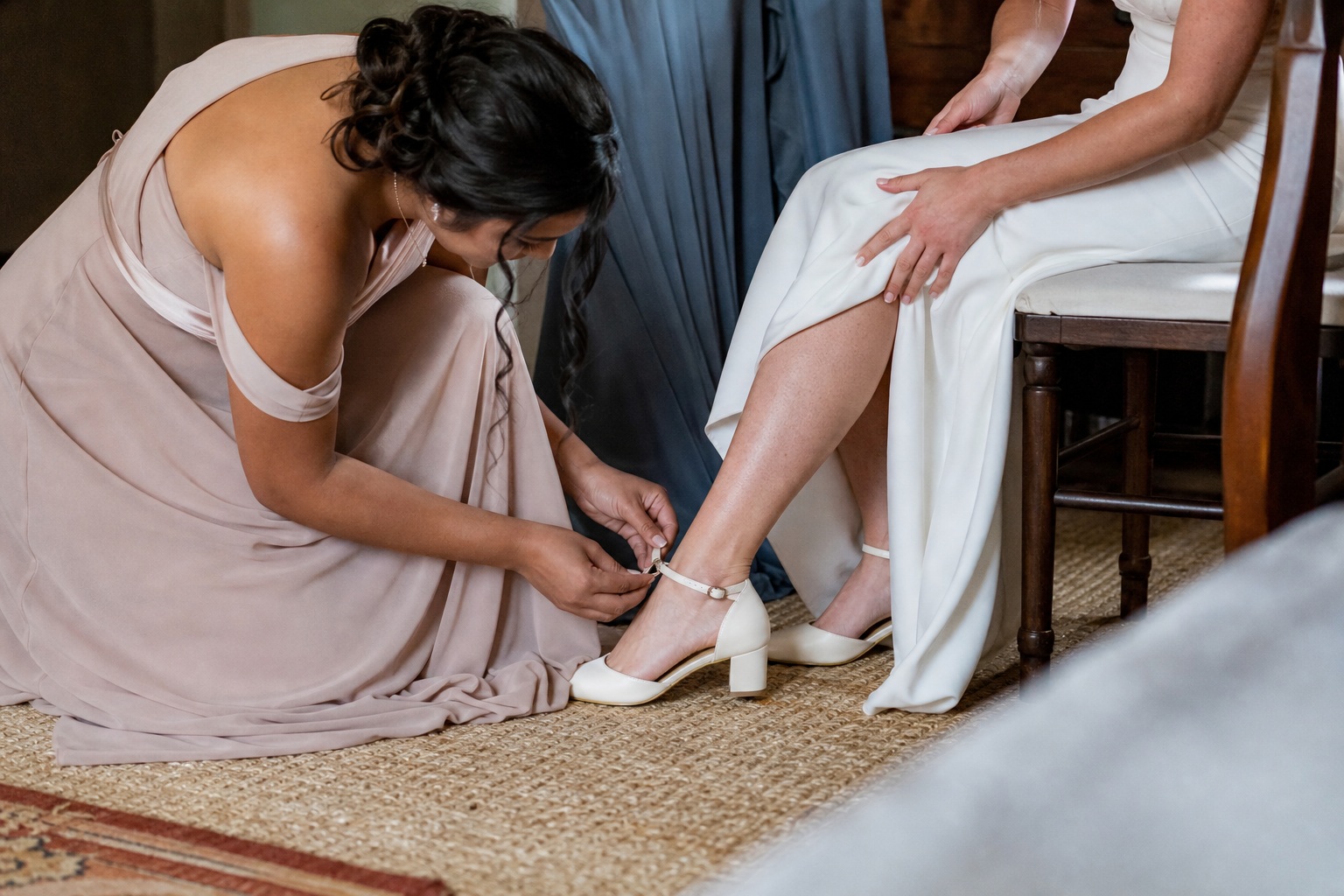 Woman kneeling to fasten a bride’s white shoe during wedding preparation