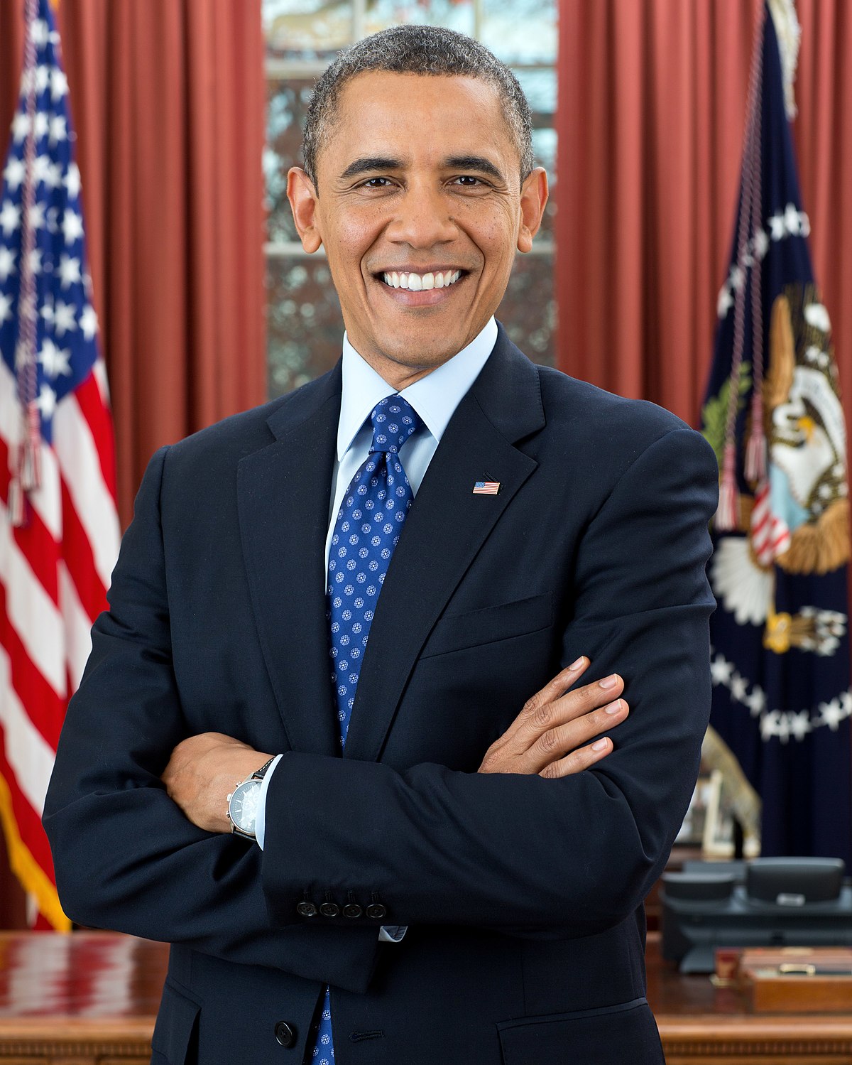 Barack Obama standing with arms crossed in the Oval Office, with flags behind him.