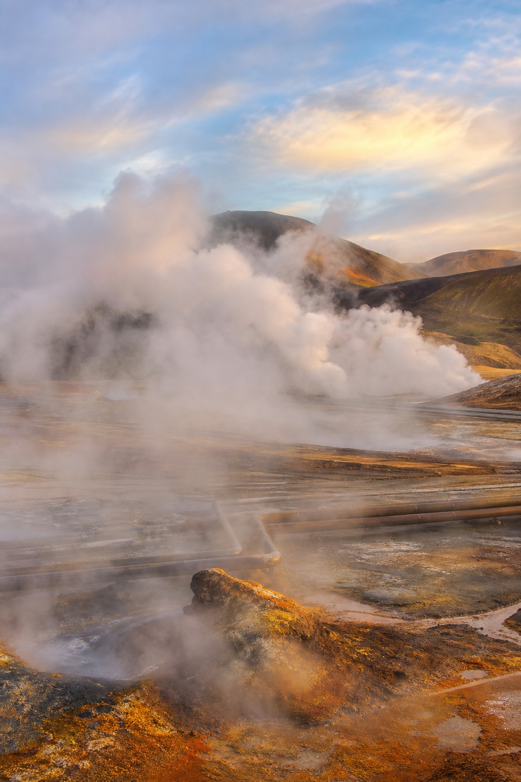 Steam rises over a volcanic geothermal landscape with rocky ground and distant hills