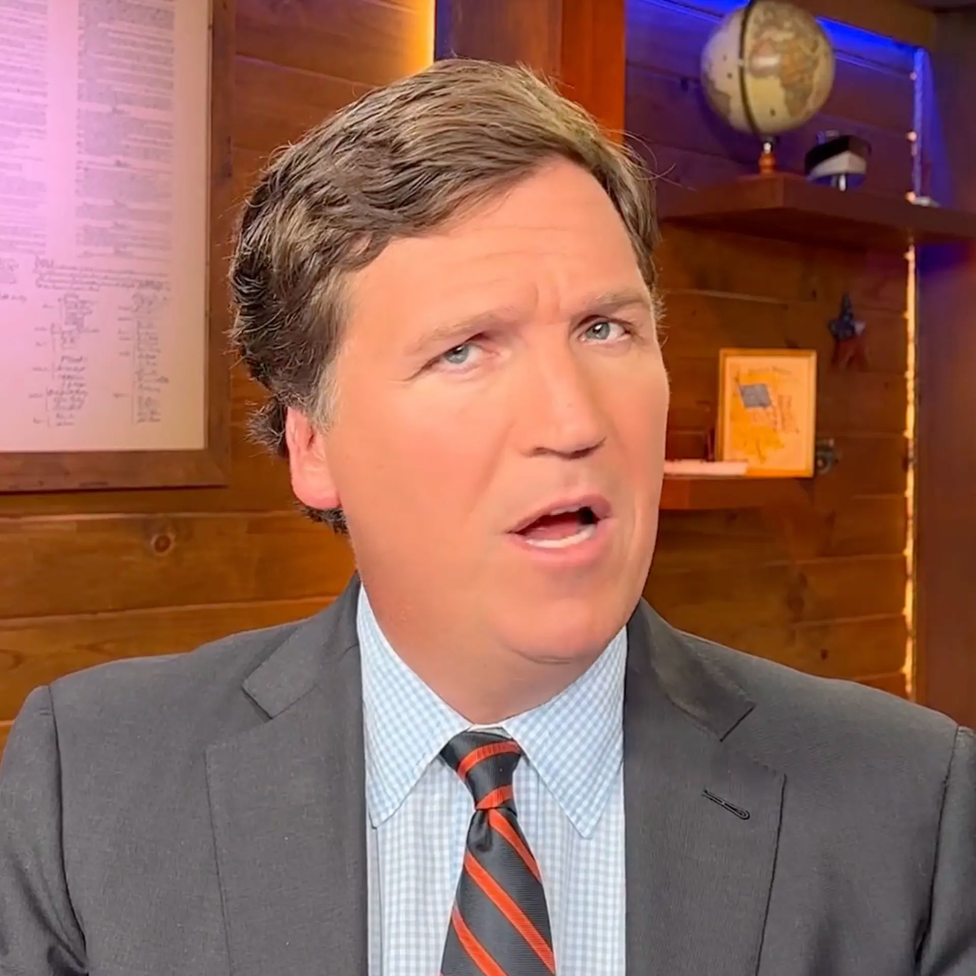 Man in a suit speaking indoors with framed posters on the wall.