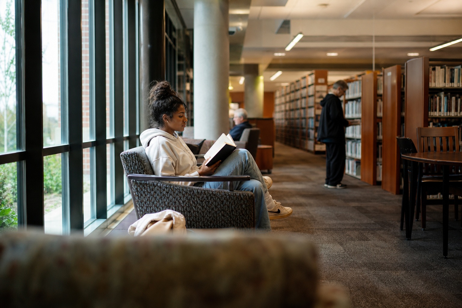 Person reading in a chair by the window inside a quiet public library.