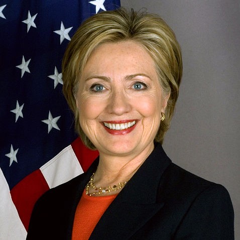 Hillary Clinton smiling in an official portrait with an American flag background.