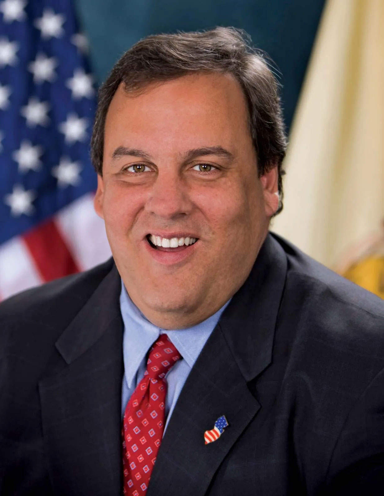 Chris Christie smiling in a dark suit, red tie, and flag pin with flags behind him