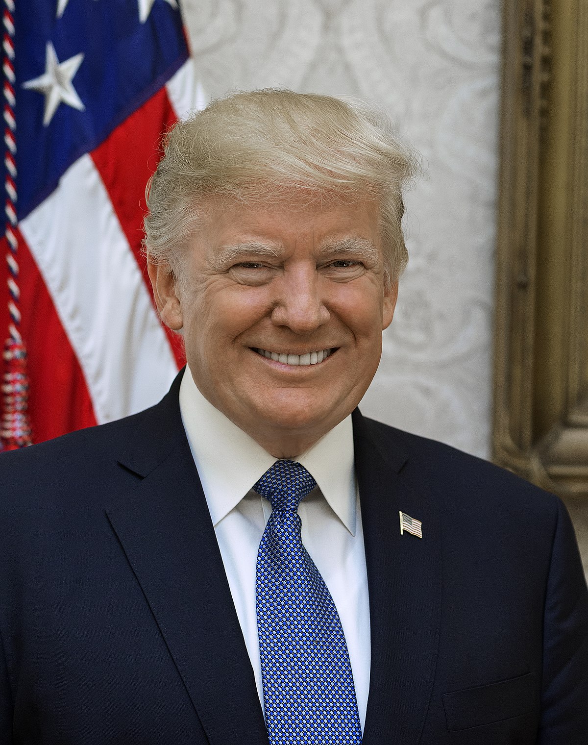 Smiling man in a dark suit and blue tie standing before an American flag.