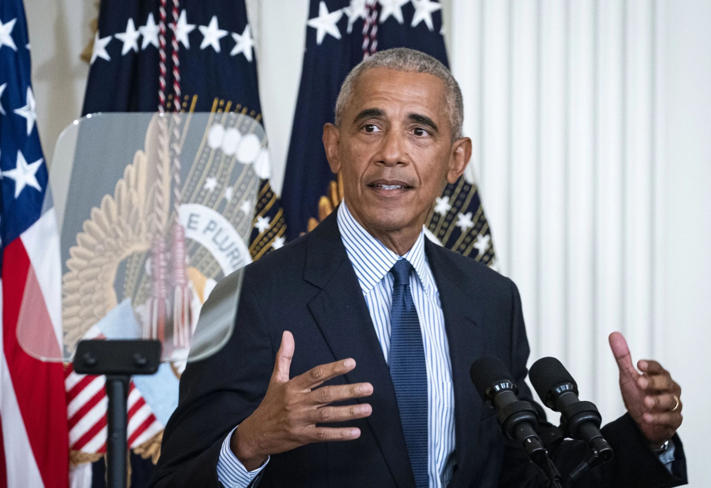 Barack Obama speaking at a podium with microphones in front of U.S. flags and the presidential seal