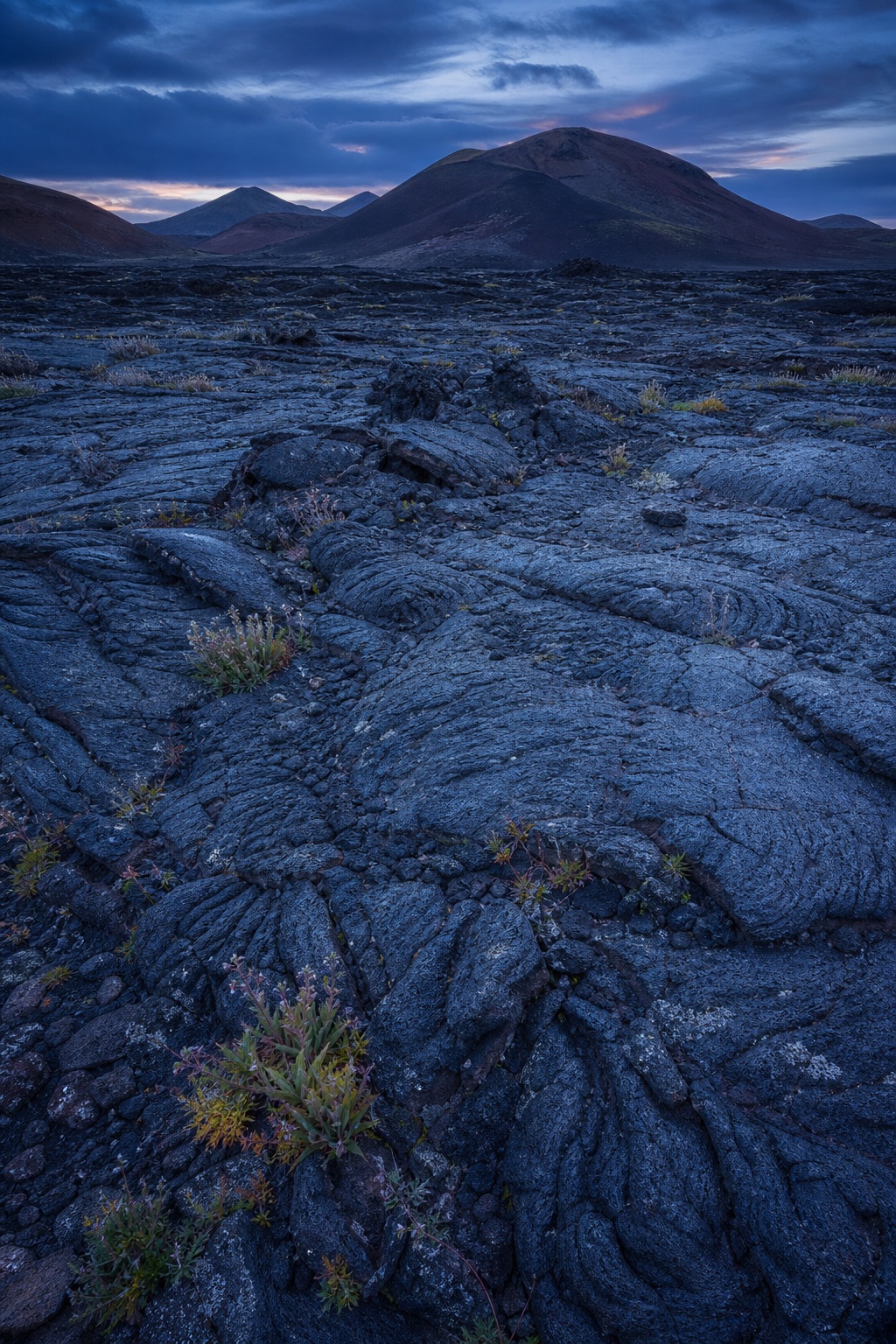 Dark cooled lava field with rugged basalt texture under a blue hour sky and distant volcanic hills