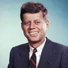 Smiling John F. Kennedy portrait in a dark suit and striped tie.