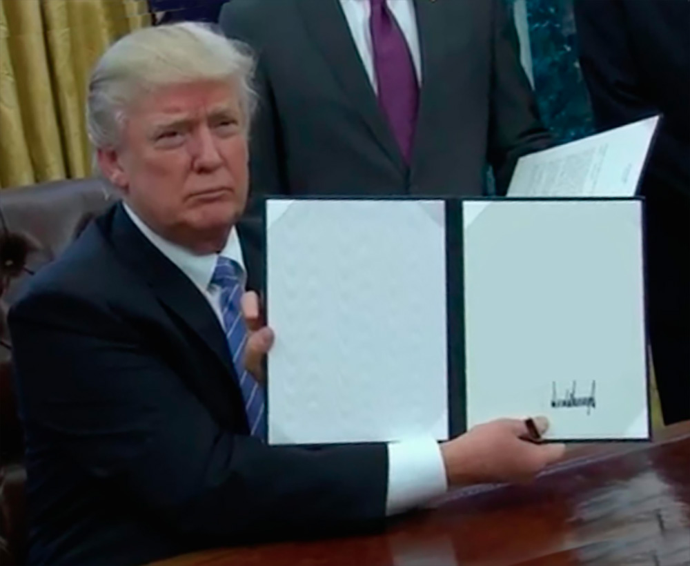 Donald Trump in a suit signing an open document at a wooden desk.