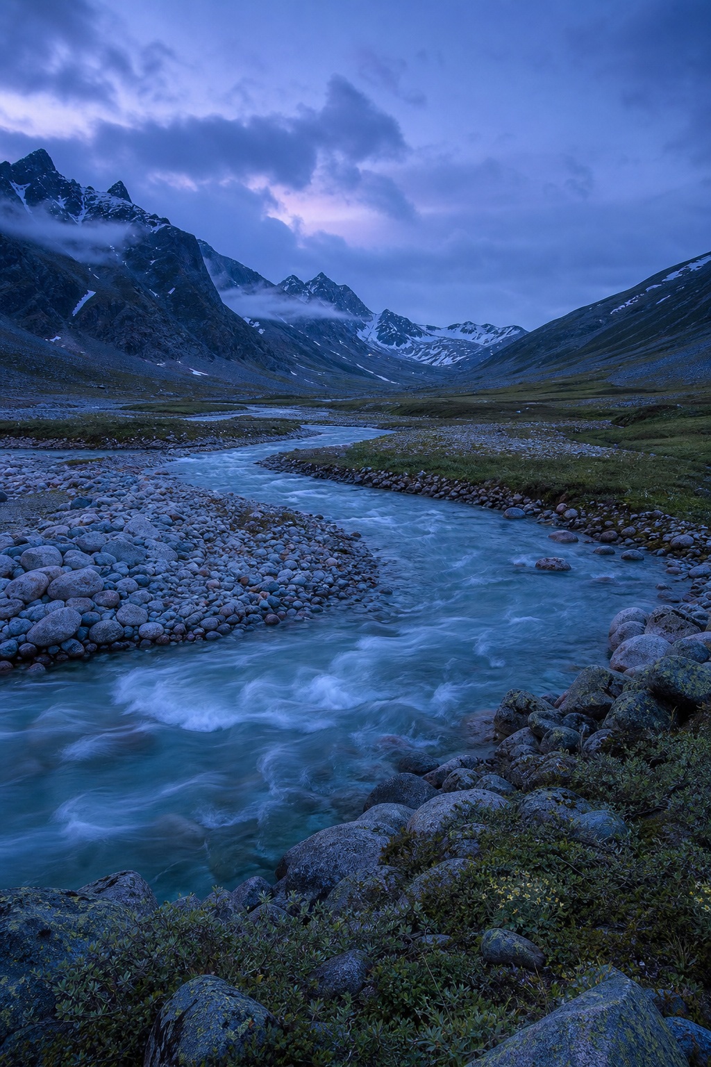 A winding blue river flows through a rocky glacial valley with mountains at dusk.