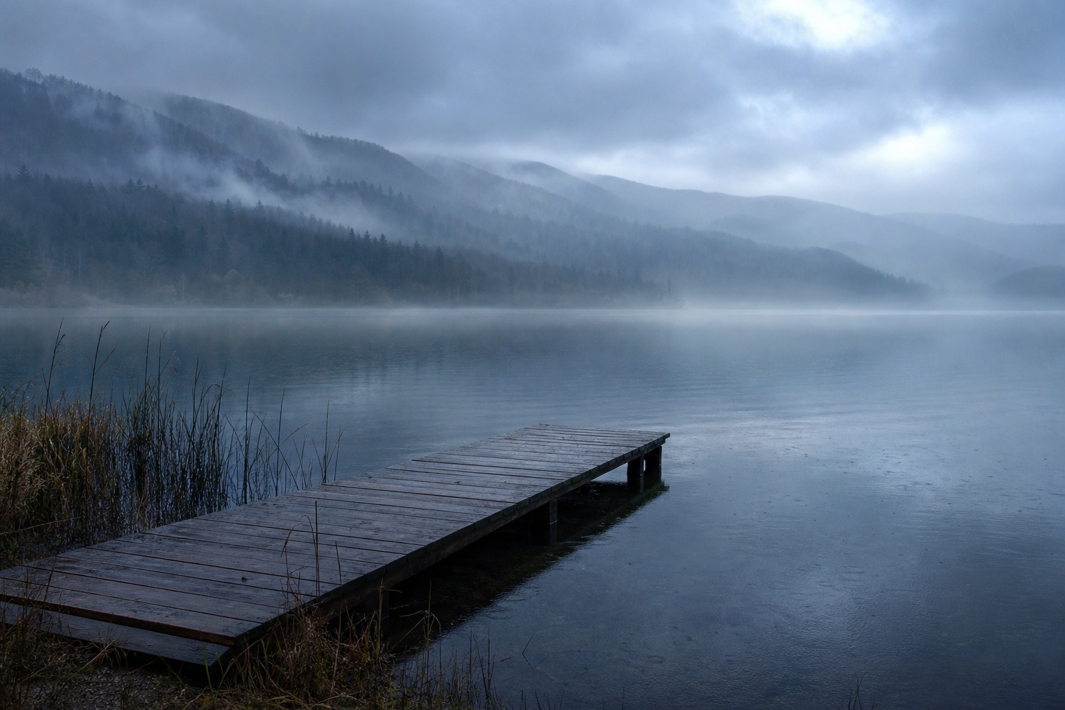 Wooden dock extending into a misty lake with mountains and reeds at dawn