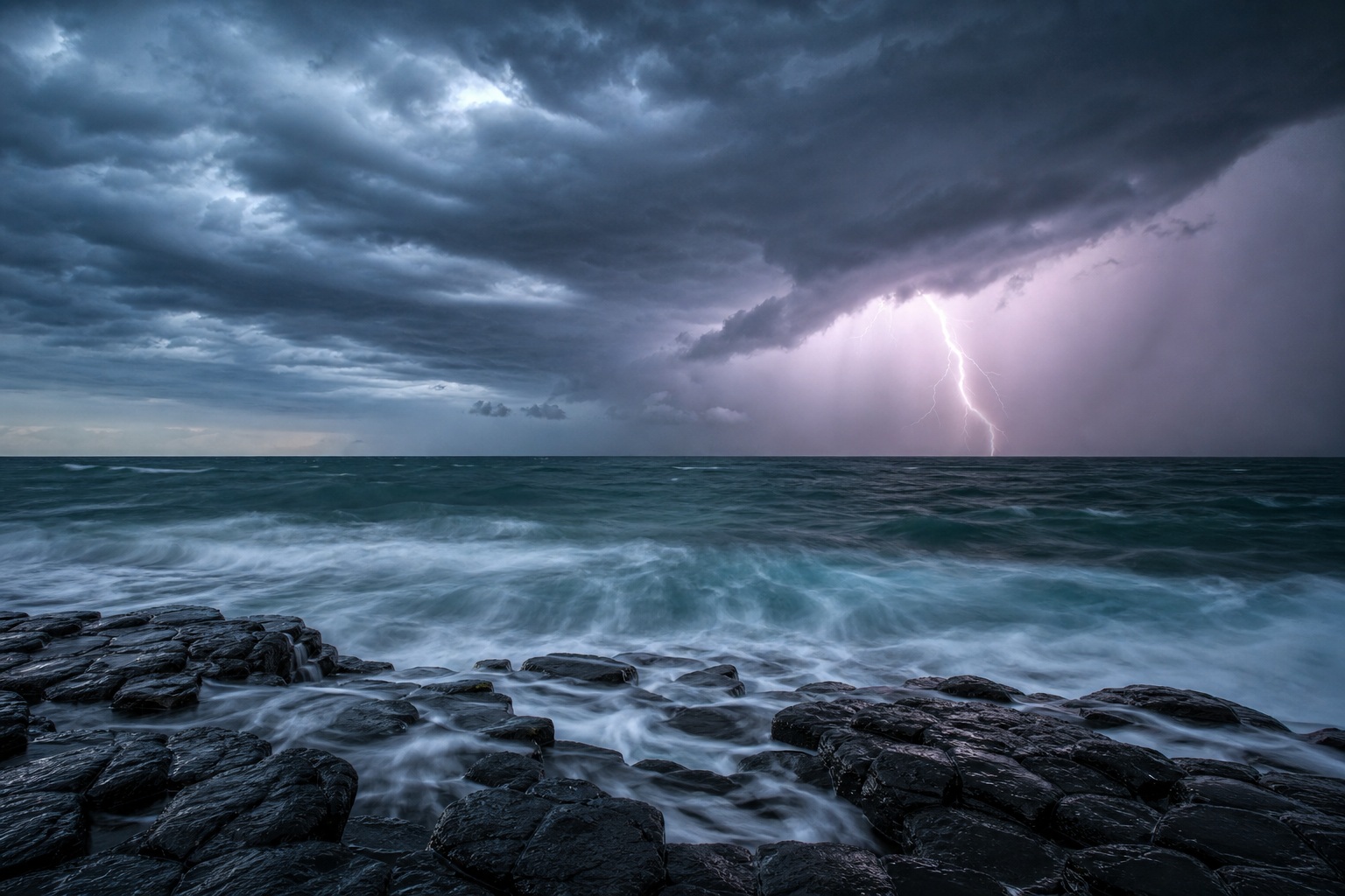 Lightning strikes over a stormy ocean beside dark basalt rocks and heavy clouds.