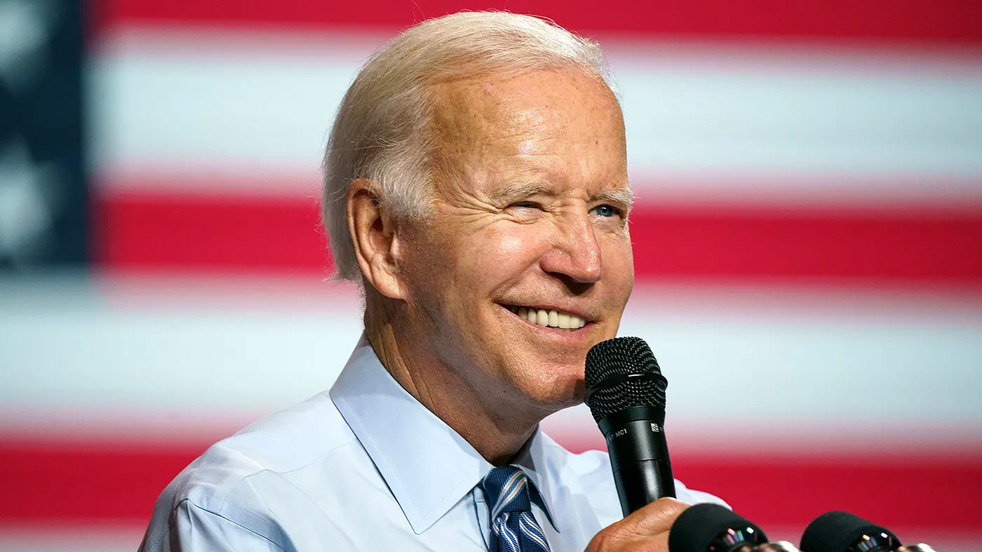Joe Biden speaking into a microphone with an American flag backdrop.