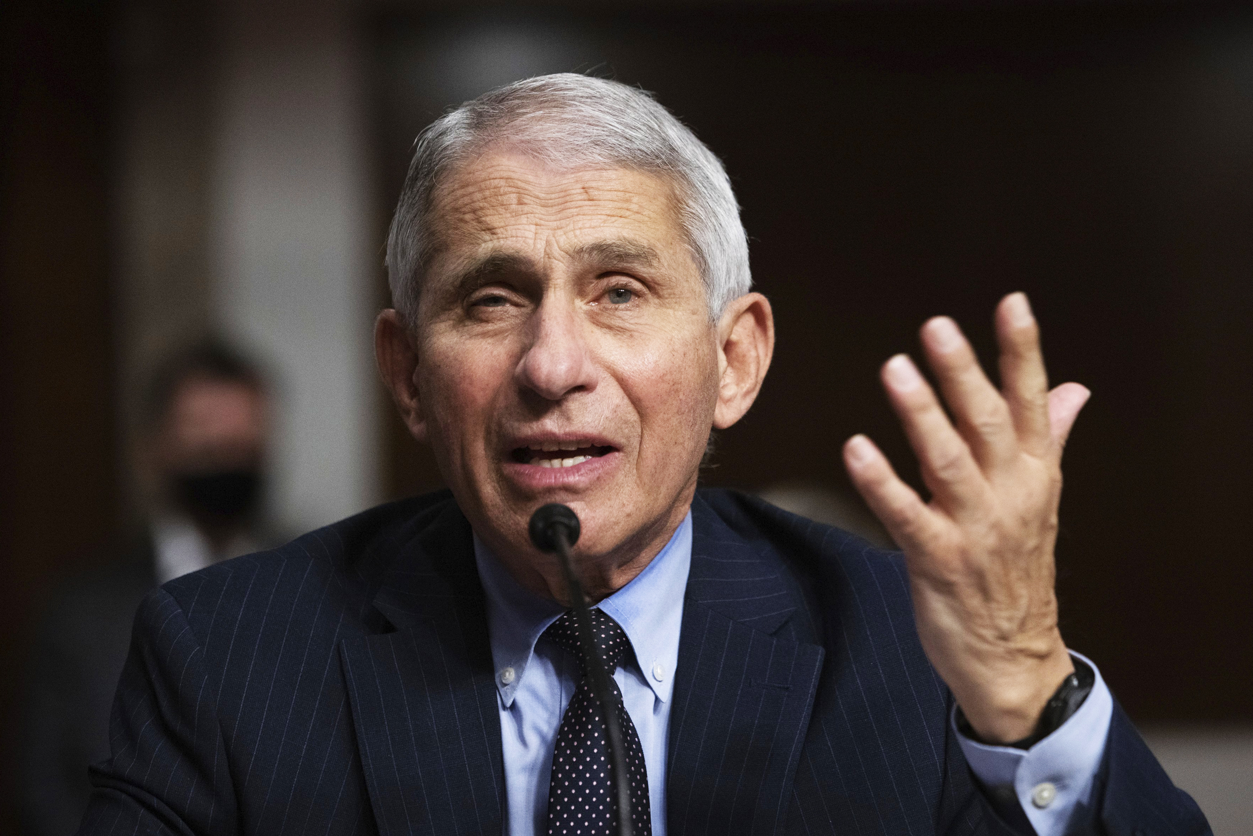 Older man speaking at a microphone with one hand raised.