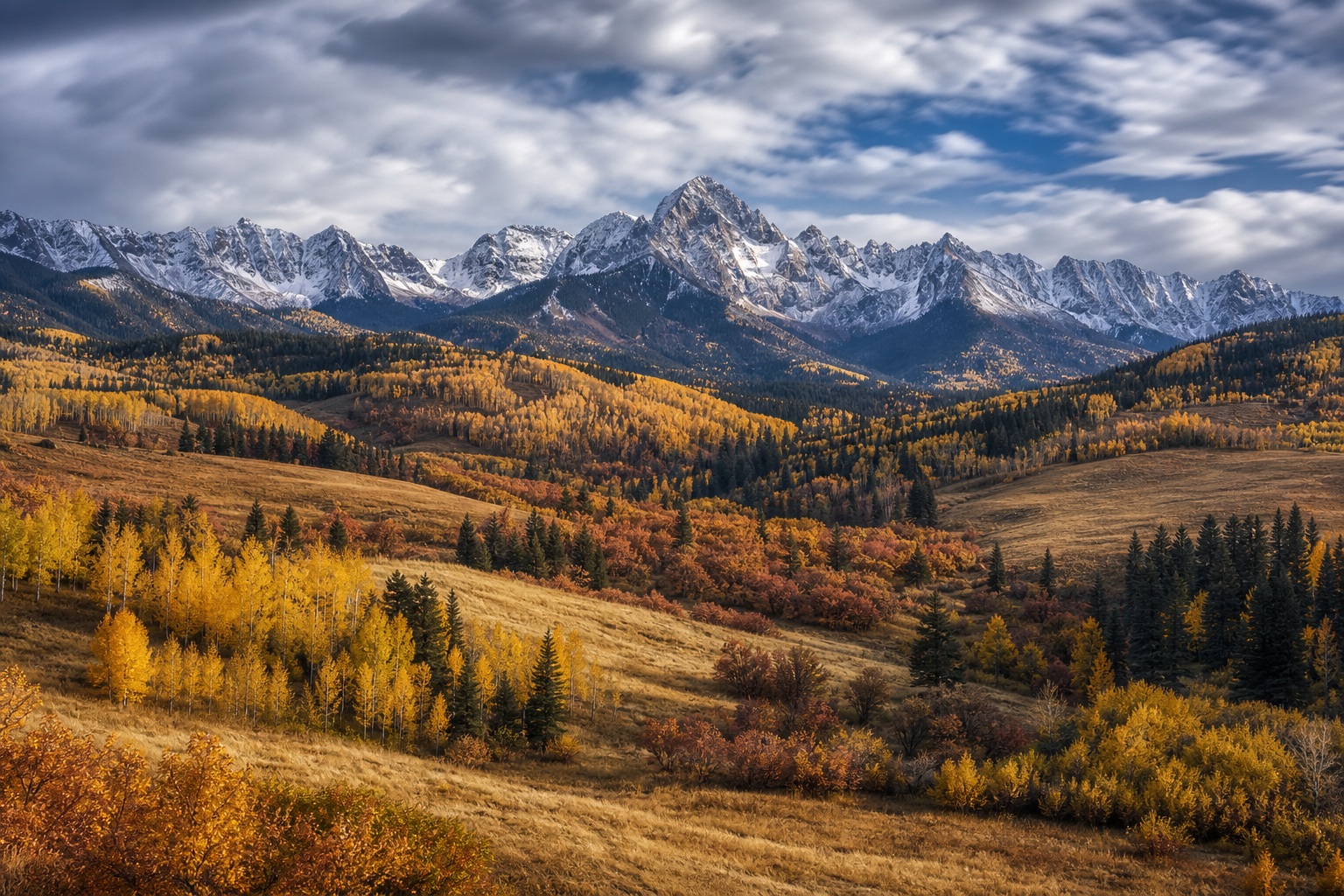 Autumn mountain valley with golden trees, snowy peaks, and rolling hills under cloudy sky