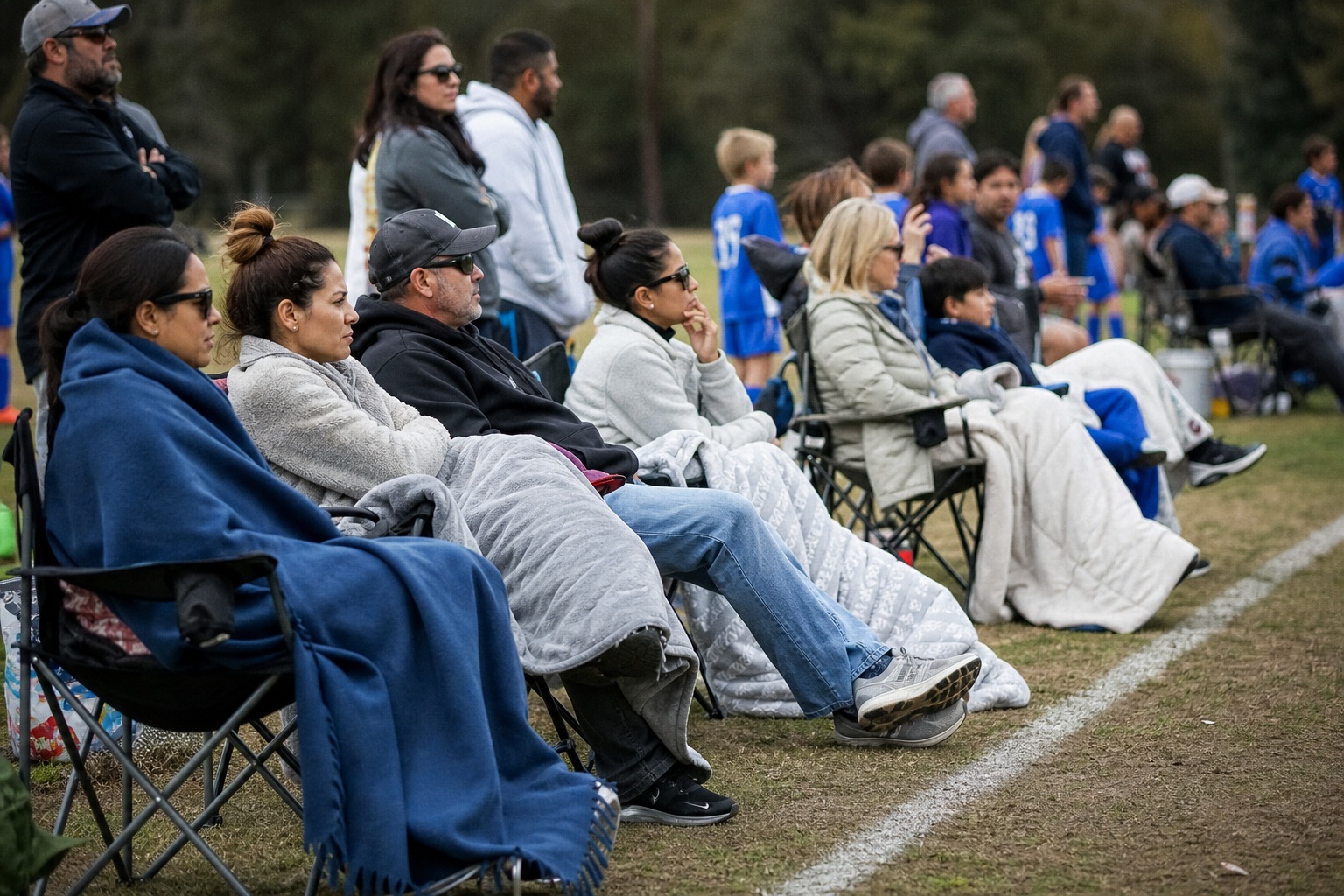 Candid fans seated in folding chairs along a soccer sideline during a youth match.