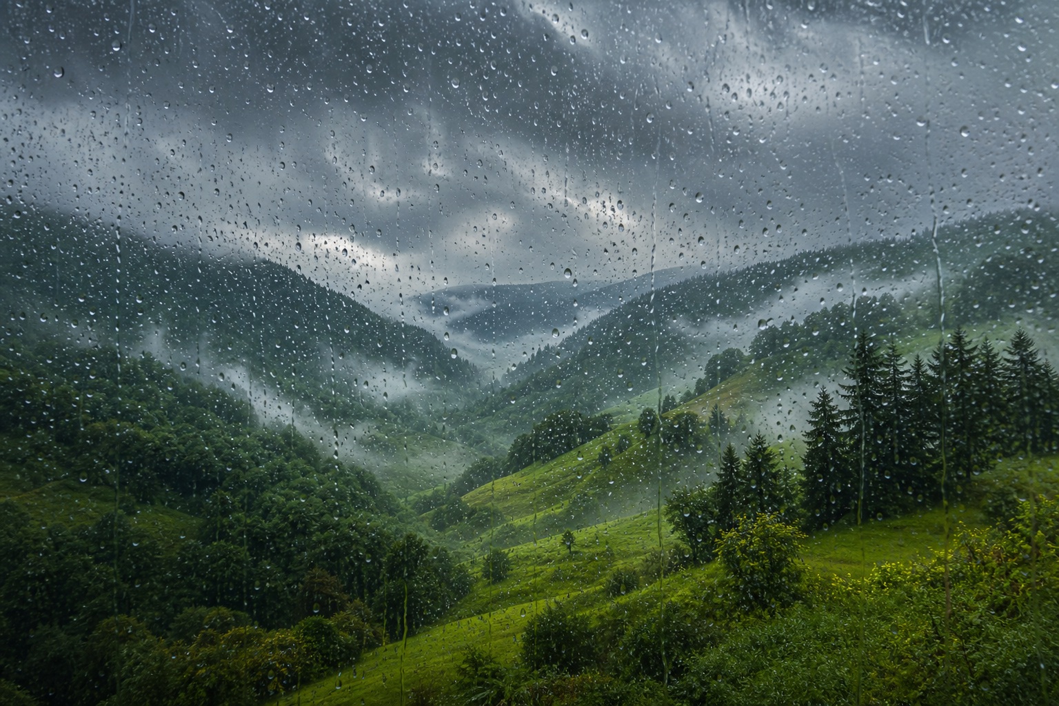 Rain-speckled window view of a misty green valley and forested hills