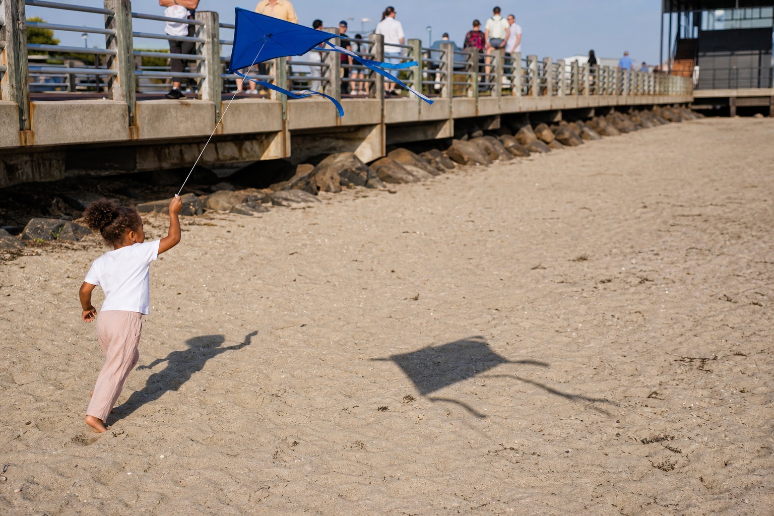 People on a weathered beach boardwalk above sand with a child flying a kite below.