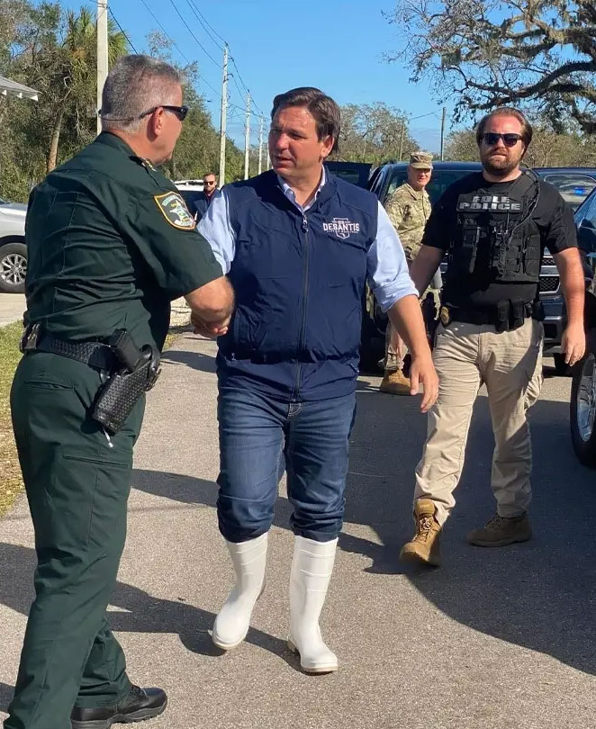 Man being escorted by a law enforcement officer while another officer stands nearby outdoors.