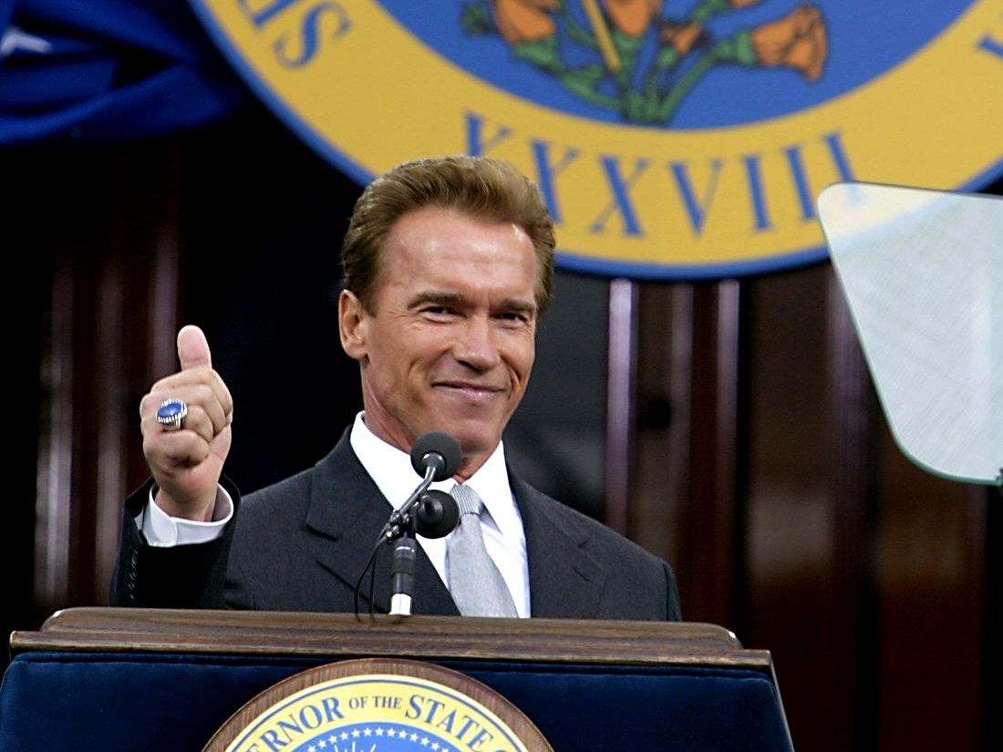 Arnold Schwarzenegger speaking at a podium and giving a thumbs-up gesture.