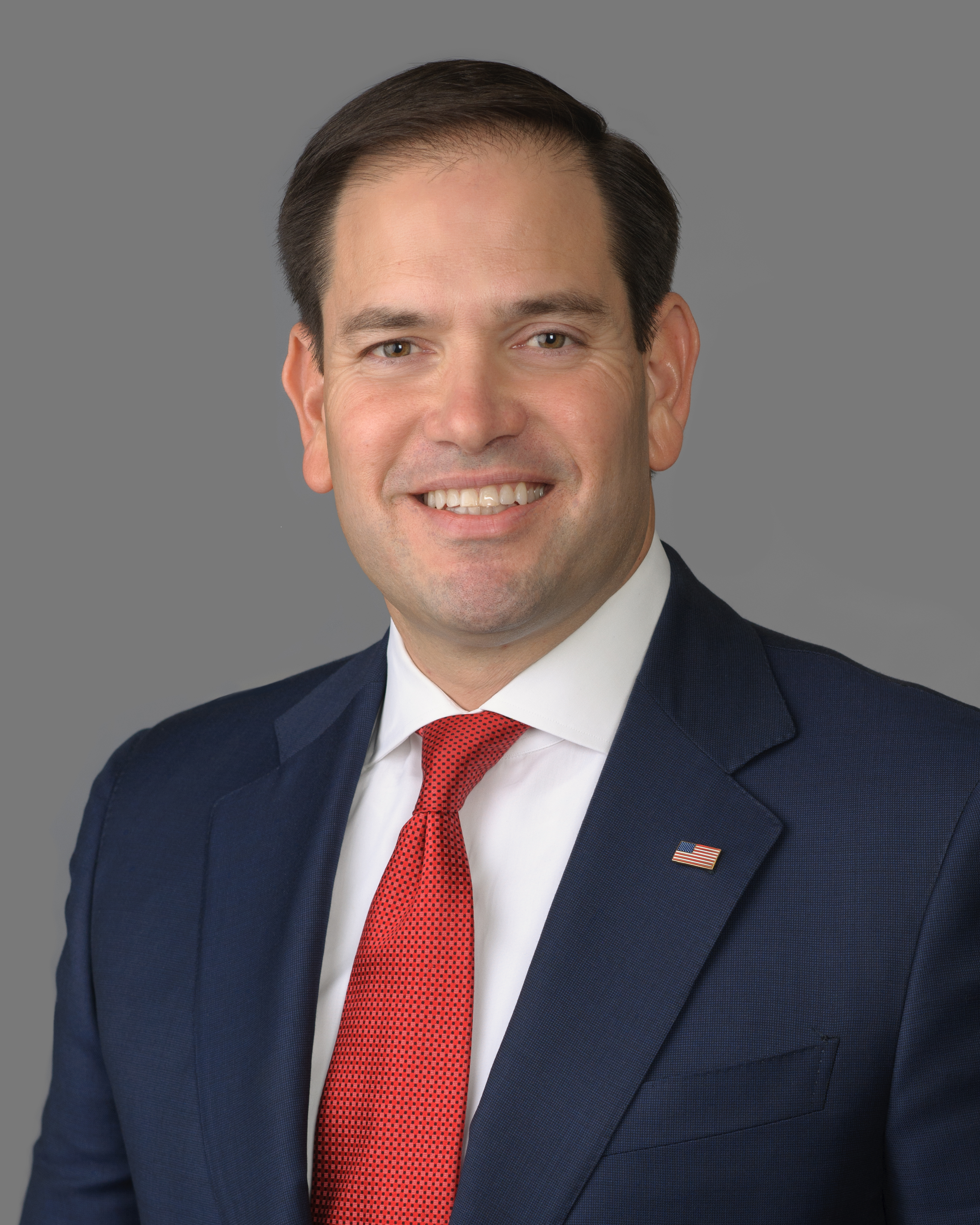 Smiling man in a navy suit and red tie against a gray background