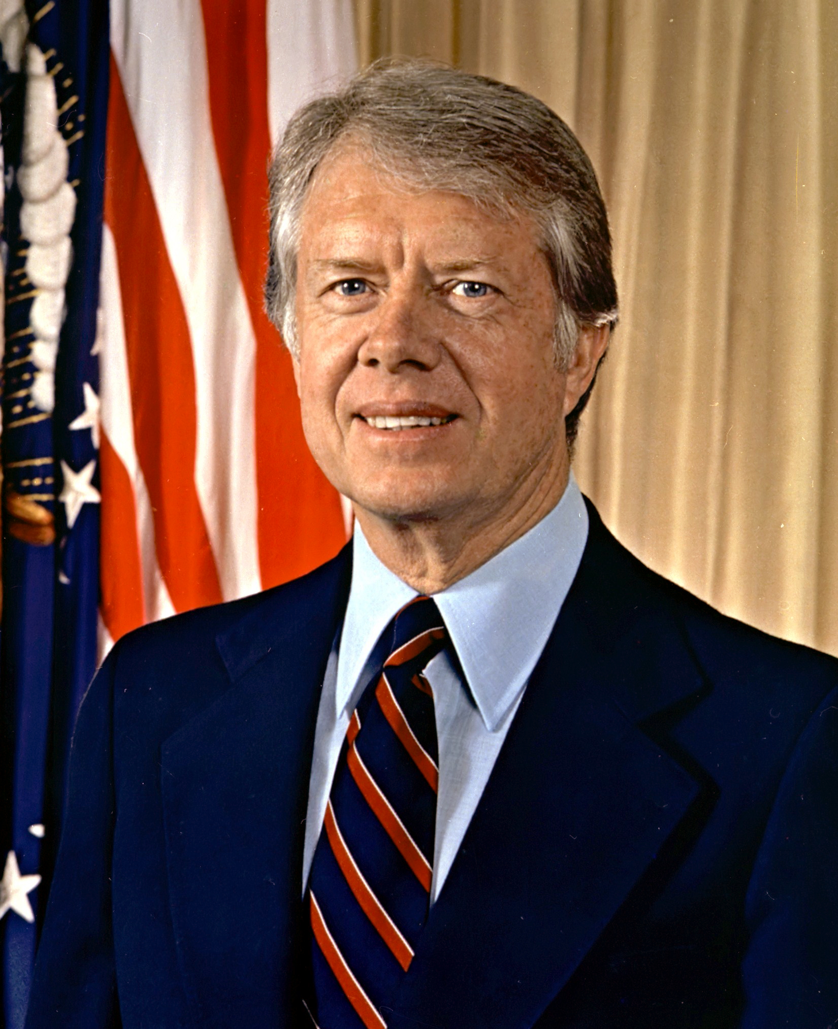 Jimmy Carter in a dark suit and striped tie standing before flags and drapery.