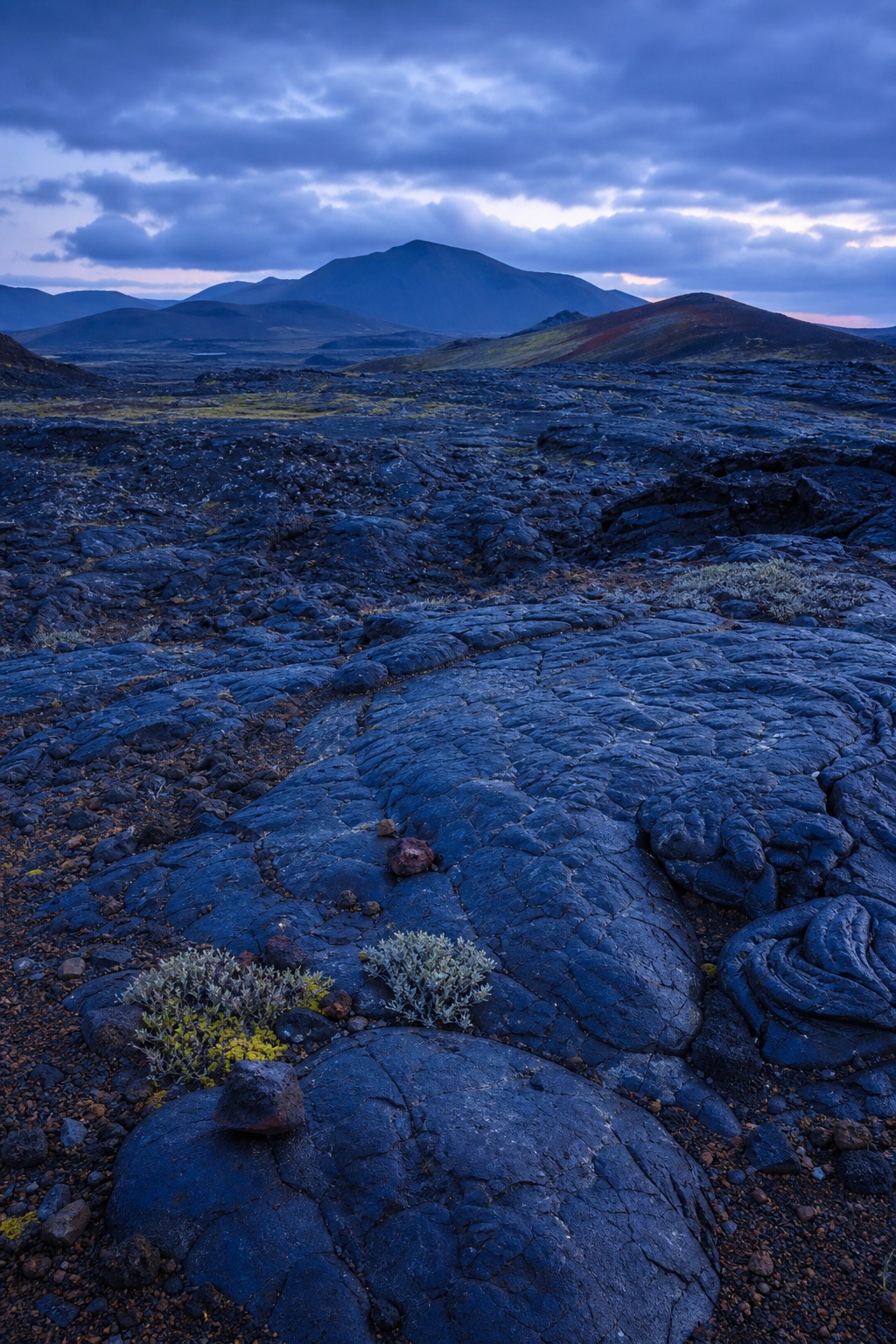 Rugged basalt lava field with distant mountains under blue hour clouds.