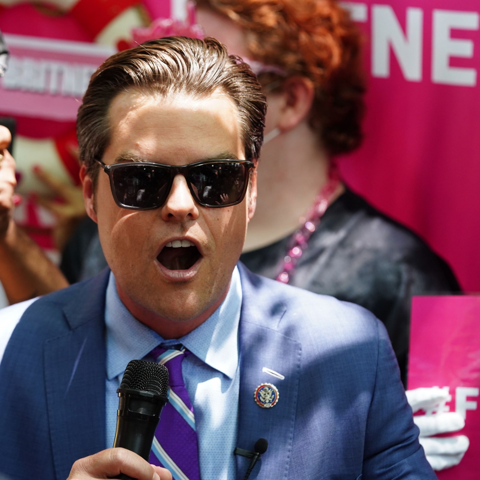 Man in suit wearing sunglasses speaking into a microphone at a rally.
