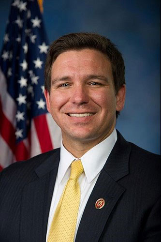 Smiling man in a suit and yellow tie in front of a U.S. flag backdrop.