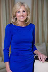 Smiling woman in a bright blue dress standing beside a chair in an indoor setting.
