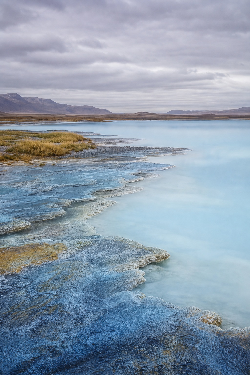 Blue mineral shelf beside pale water with distant mountains under cloudy sky.