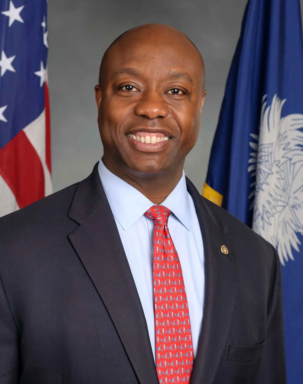 Smiling man in a dark suit and red tie standing before flags.