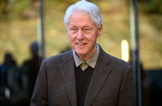 An elderly man wearing a dark suit and tie, standing outdoors with a blurred background.