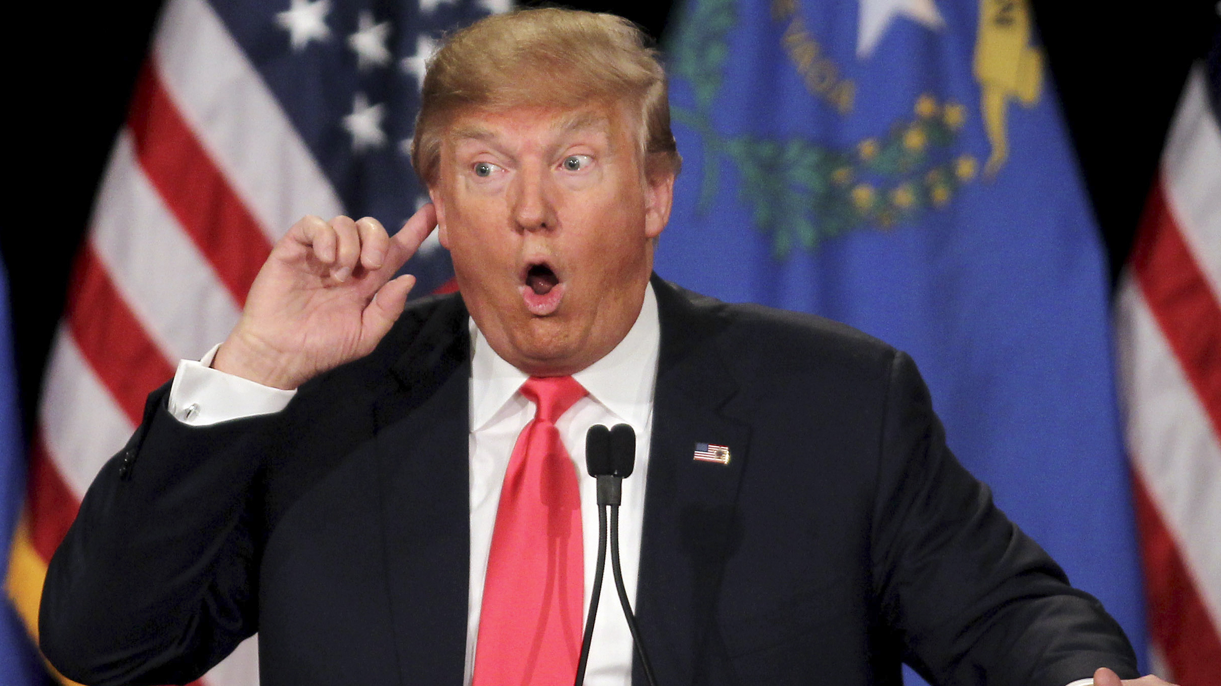 Donald Trump speaking at a rally with flags in the background and one hand raised.