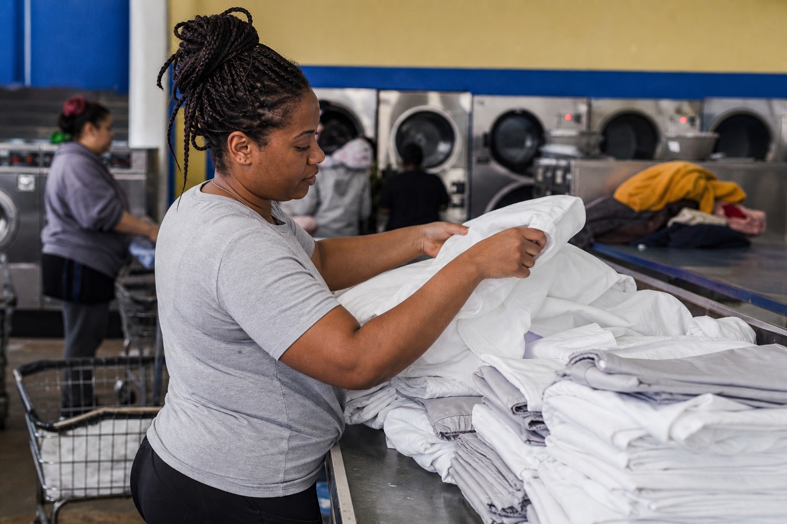 Woman folding white linens beside stacked towels in a laundromat.