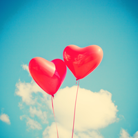 Two red heart-shaped balloons floating in the air against a blue sky with white clouds
