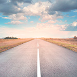 Empty road with fields on either side under a cloudy blue sky