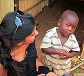 A woman looking at a young boy in a striped shirt, sitting in a rural area with a dirt floor and wooden structures.
