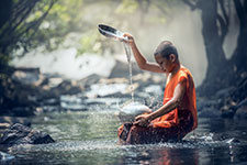 Monk in orange robes pouring water from a bowl into a forest stream