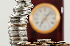 A tall, unstable stack of silver coins next to a clock face with Roman numerals on a dark red background