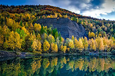 A calm lake reflecting the vibrant yellow and green trees on its banks and a large rocky hill in the background under a partly cloudy sky.
