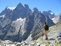 A person with a backpack and trekking poles stands on a rocky outcropping, looking out at a rugged mountain landscape.