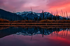A calm lake mirrors the surrounding mountains and trees under a pink and purple sky.