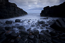 A rocky shoreline with waves crashing against dark stones between two large rock formations under a cloudy sky.