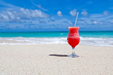 A red cocktail in a glass with a straw on a sandy beach with the ocean in the background