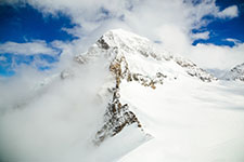 A snowy mountain peak partially obscured by clouds against a blue sky.