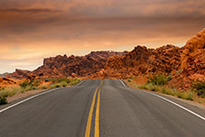 A long, straight desert road with double yellow lines, surrounded by rocky mountains and greenery, under a cloudy orange sky.